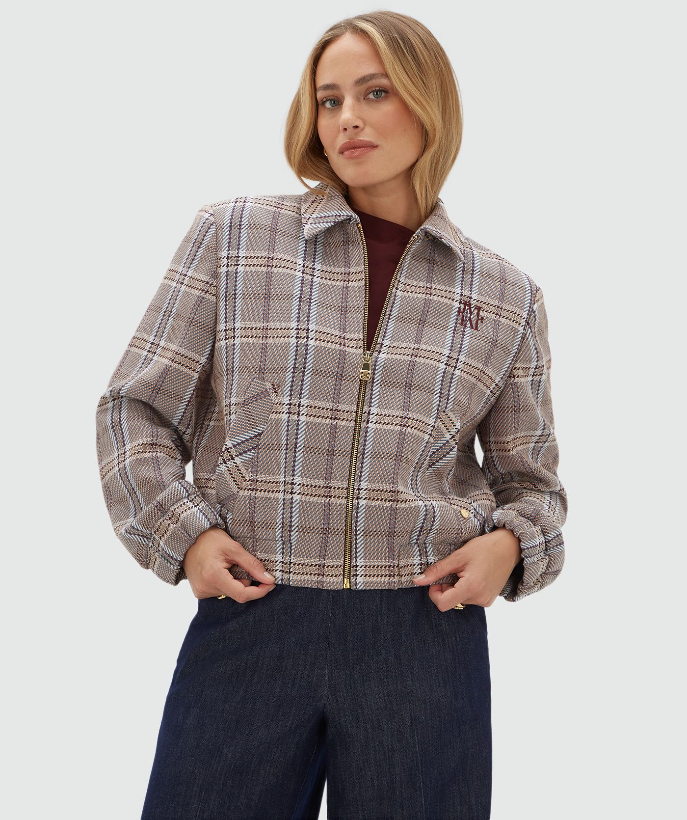 Model draagt een beige, bruin en donkerrood geruit jasje met rits gecombineerd met een donkerblauwe pantalon en een rood topje.