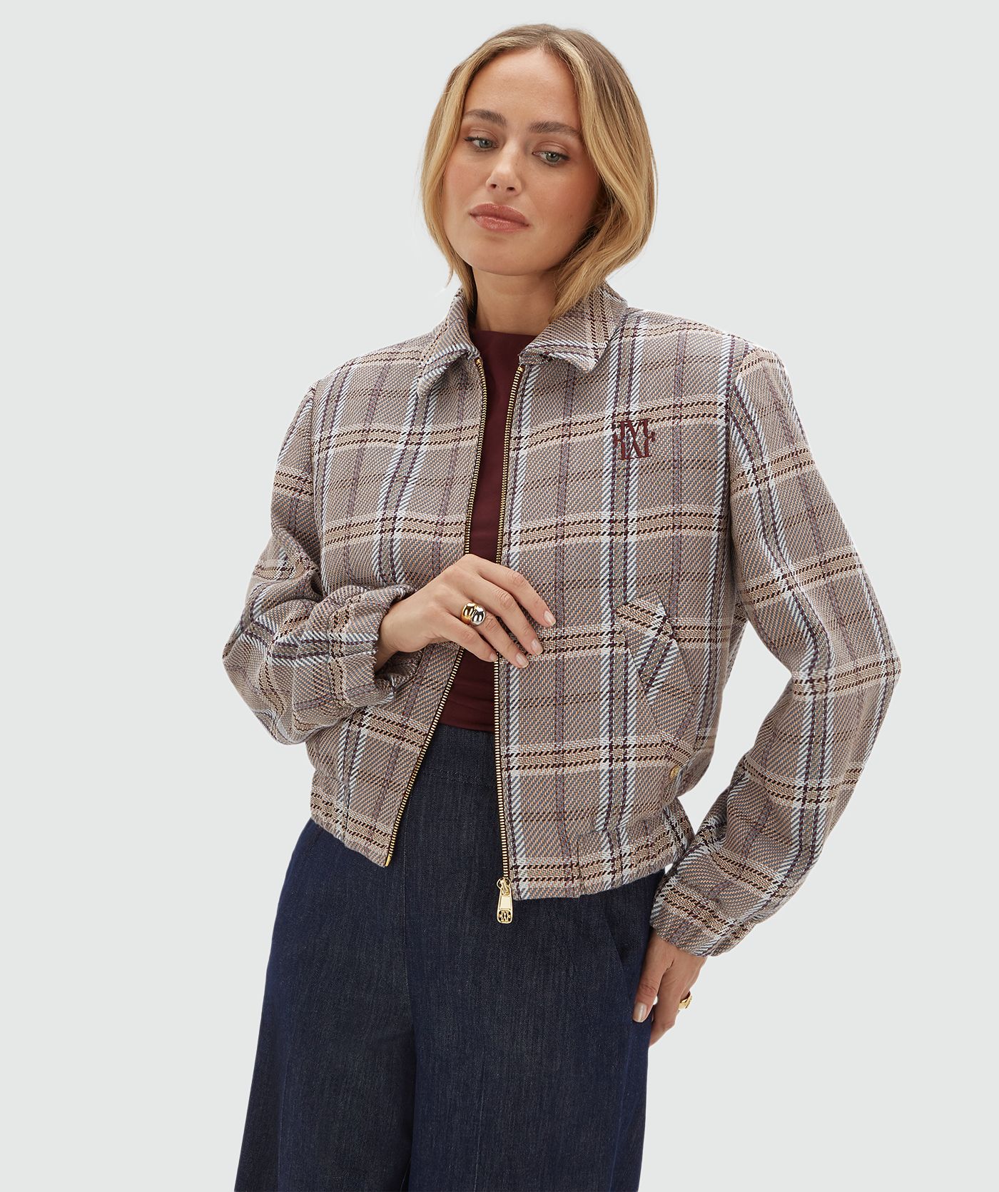 Model draagt een beige, bruin en donkerrood geruit jasje met rits gecombineerd met een donkerblauwe pantalon en een rood topje.