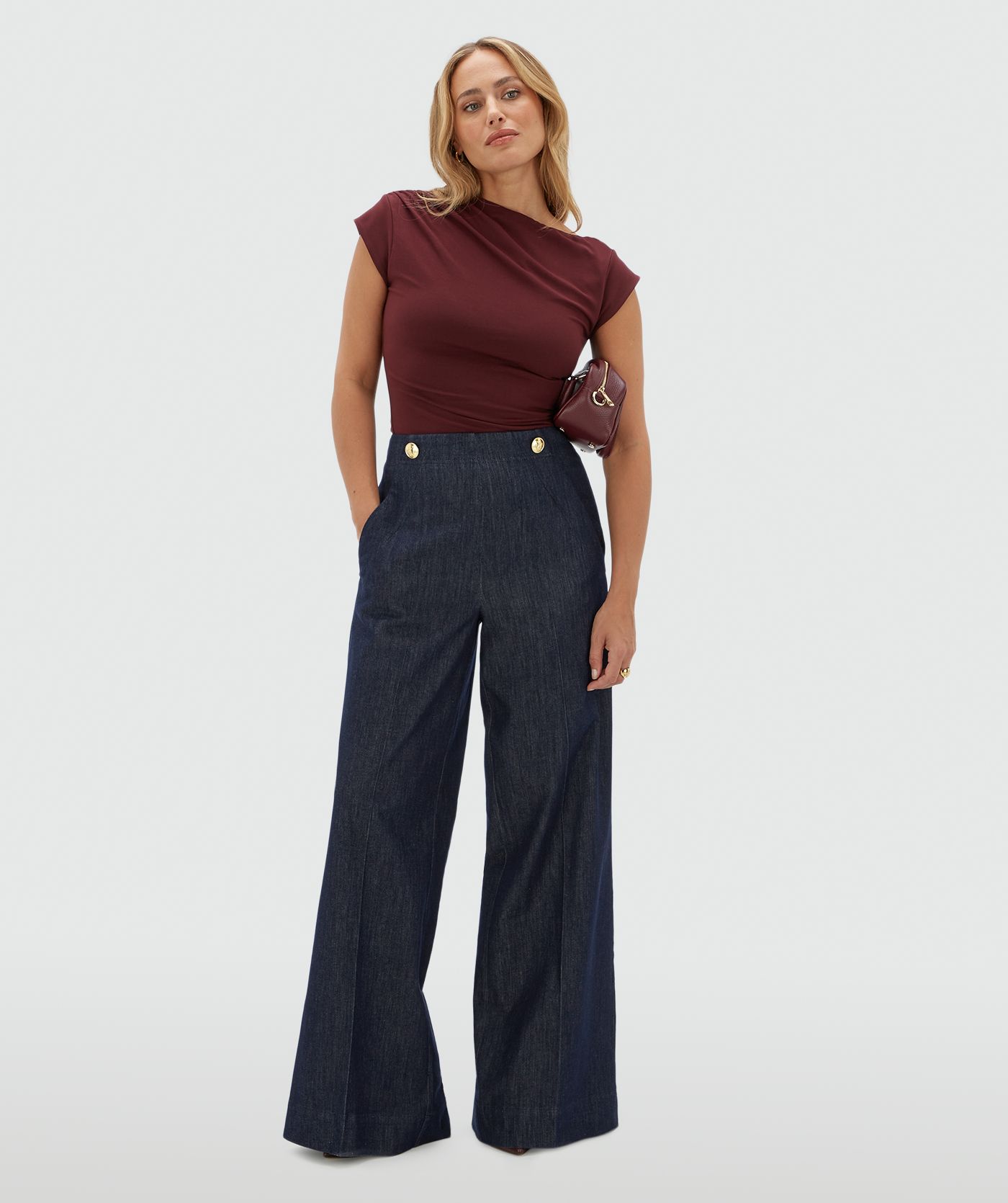Model draagt een donkerrood topje met korte mouwen en een a-symmetrische halslijn. Gecombineerd met een donkerblauwe pantalon.