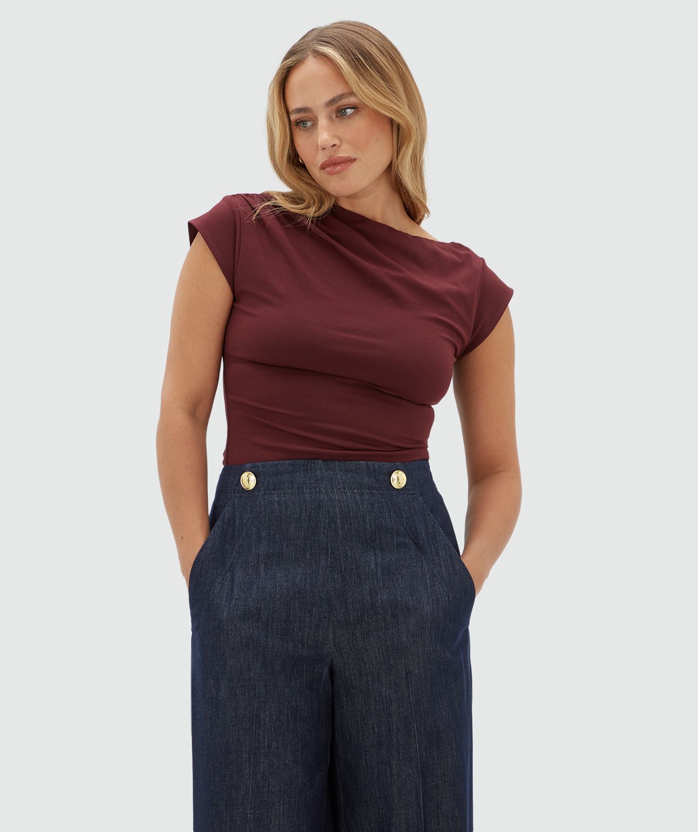 Model draagt een donkerrood topje met korte mouwen en een a-symmetrische halslijn. Gecombineerd met een donkerblauwe pantalon.