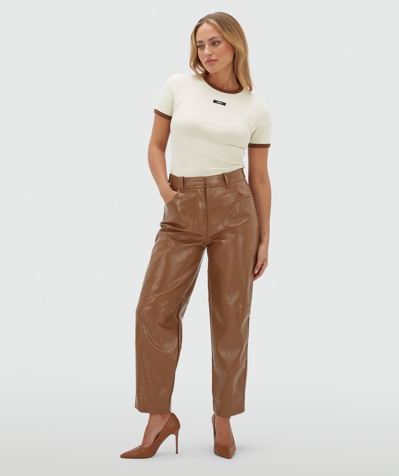 Model draagt een lichtbruine lak look broek met een wit T-shirt en heeft bruine randje bij de hals en aan het einde van de mouw.