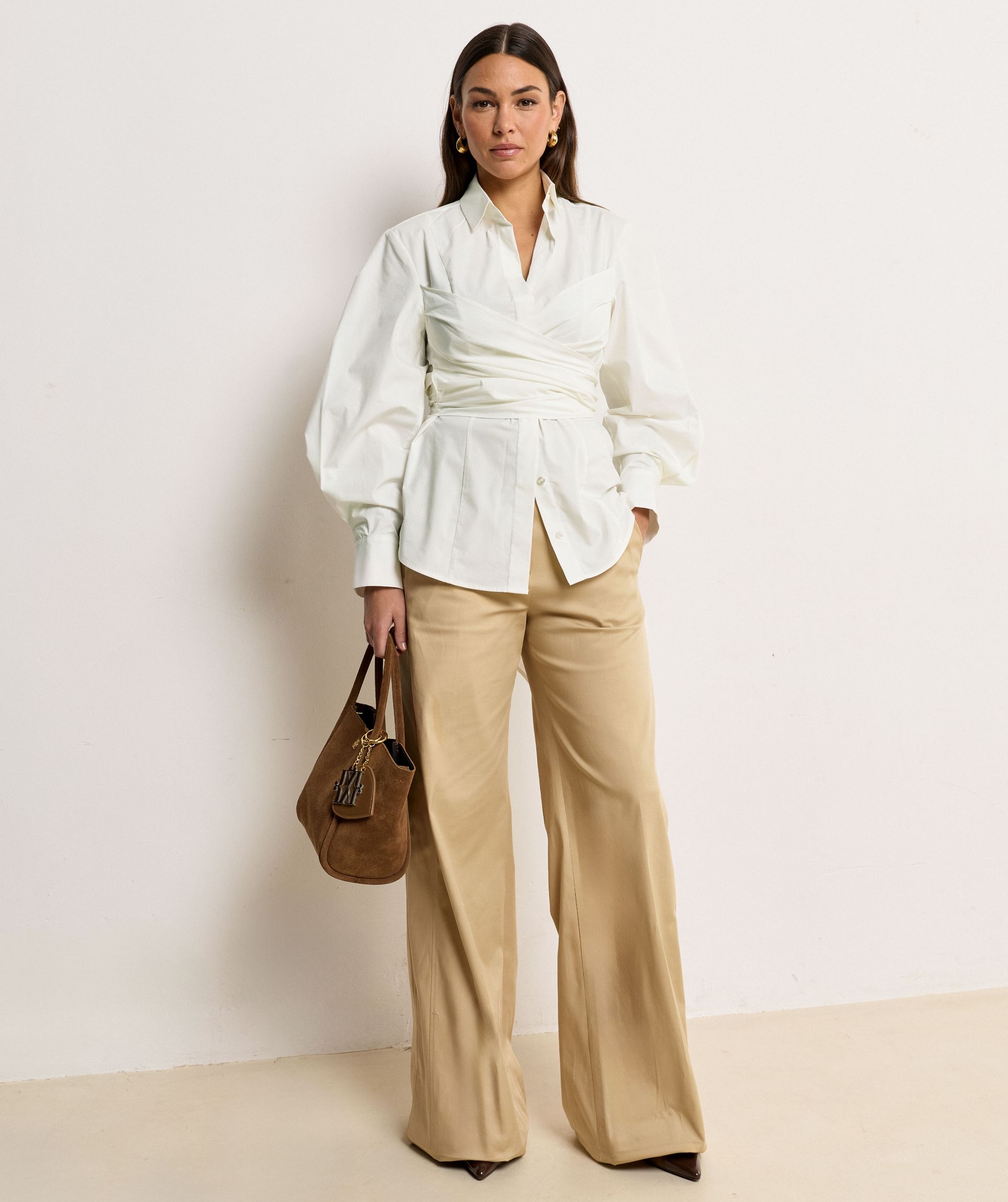 Het model draagt een mid rise wide leg pantalon in de kleur Light Camel, voorzien van verstelbare knopen in de tailleband