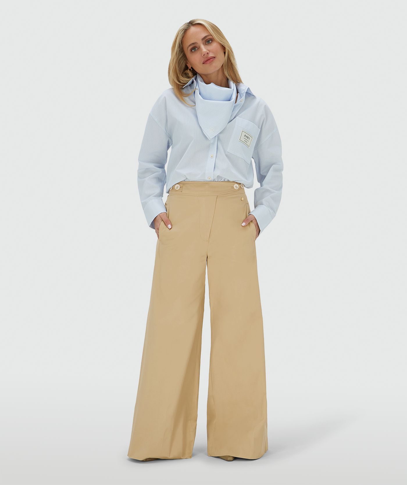 Het model draagt een mid rise wide leg pantalon in de kleur Light Camel, voorzien van verstelbare knopen in de tailleband