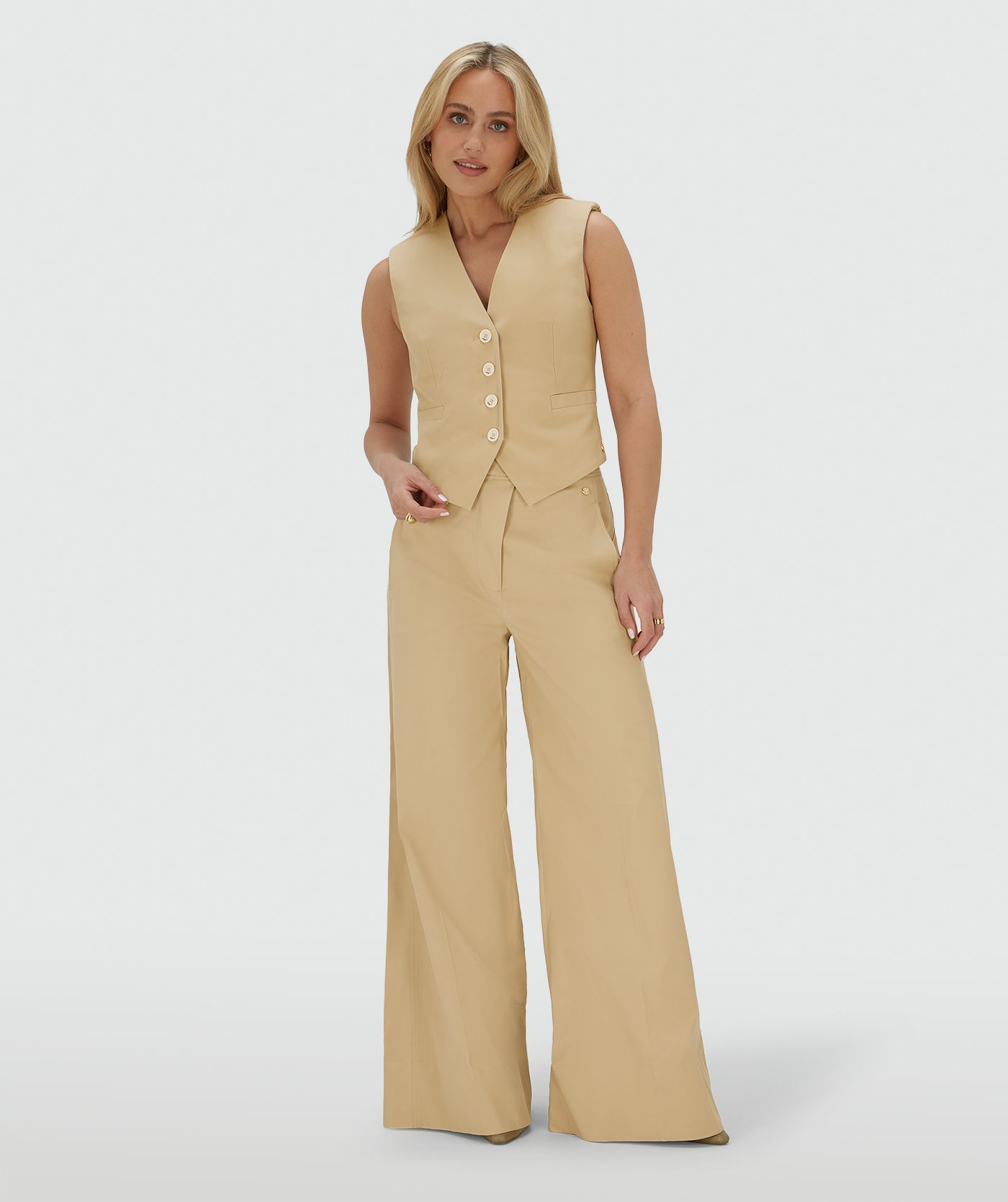 Het model draagt een mid rise wide leg pantalon in de kleur Light Camel, voorzien van verstelbare knopen in de tailleband
