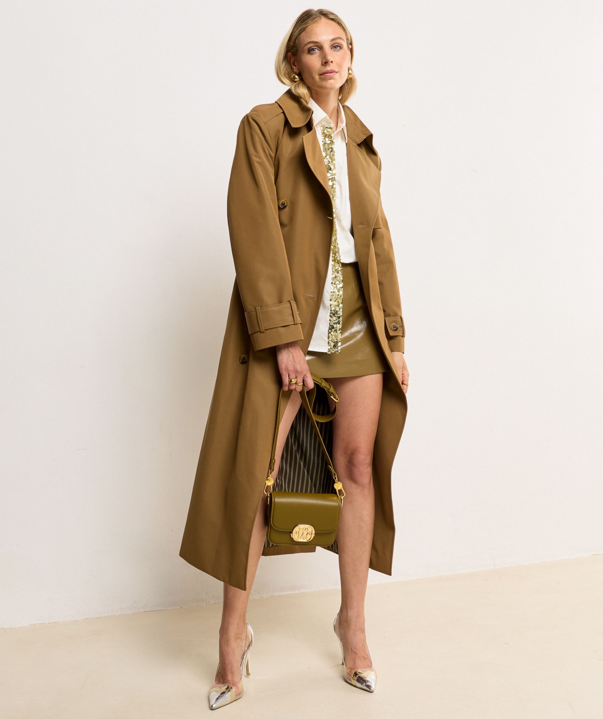 Model draagt een lange, Mid Brown trenchcoat met dubbele knopen en ceintuur in de taille