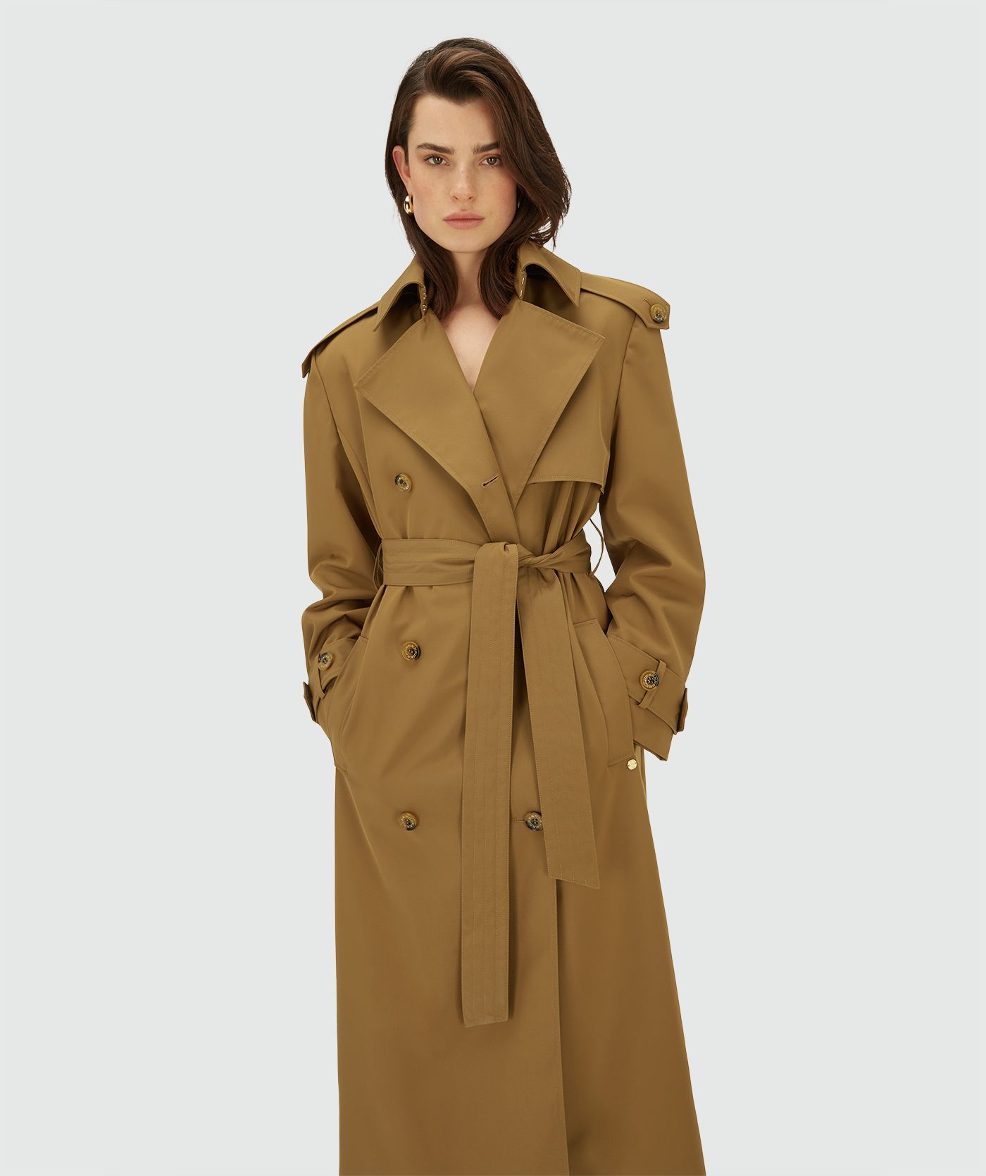 Model draagt een lange, Mid Brown trenchcoat met dubbele knopen en ceintuur in de taille