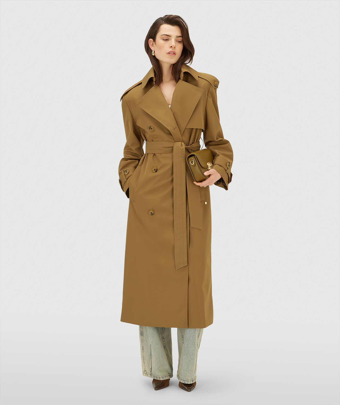 Model draagt een lange, Mid Brown trenchcoat met dubbele knopen en ceintuur in de taille
