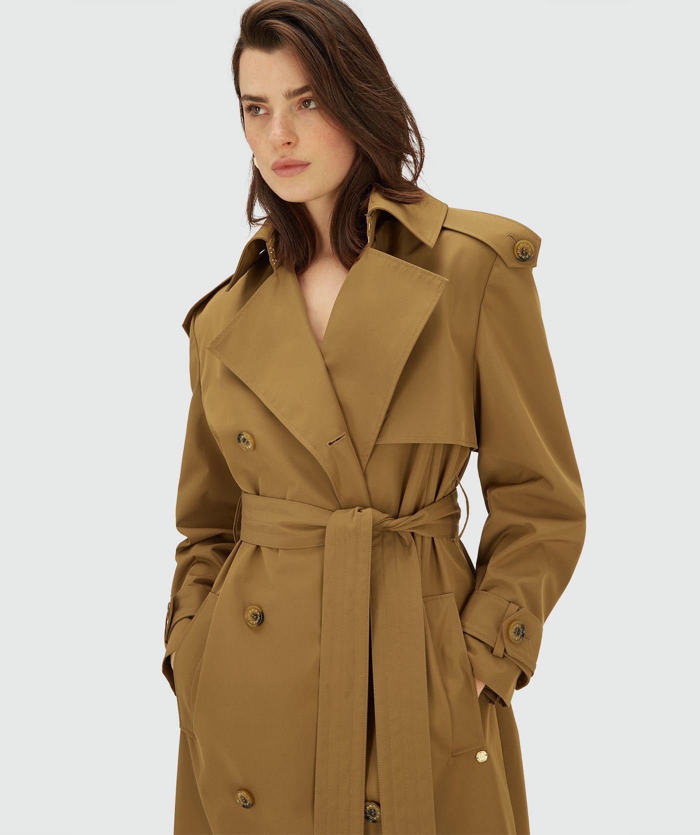 Model draagt een lange, Mid Brown trenchcoat met dubbele knopen en ceintuur in de taille