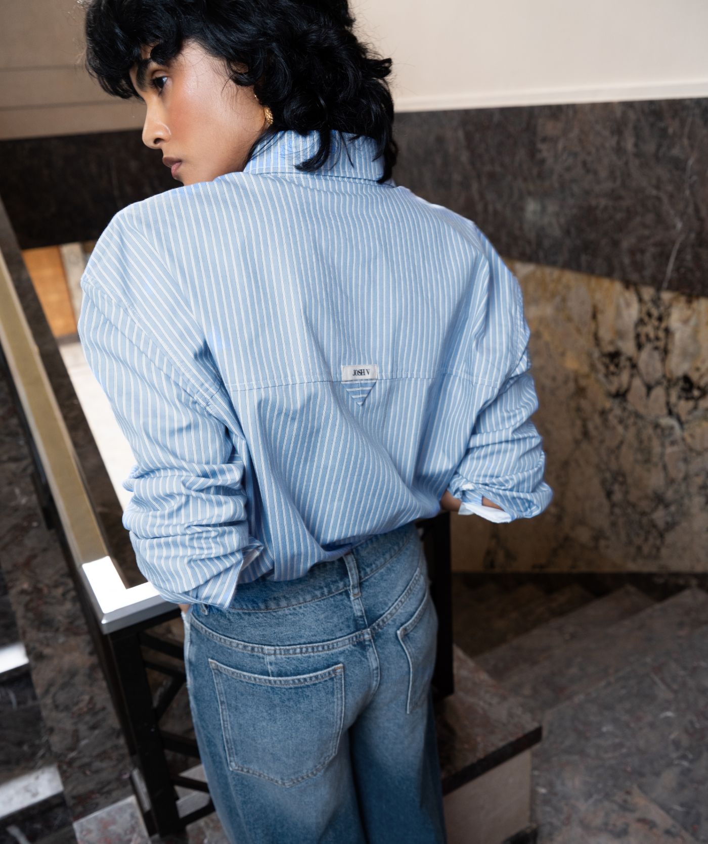 Achteraanzicht van het model in een lichtblauw met wot gestreepte blouse. De blouse is gecombineerd met een jeans.