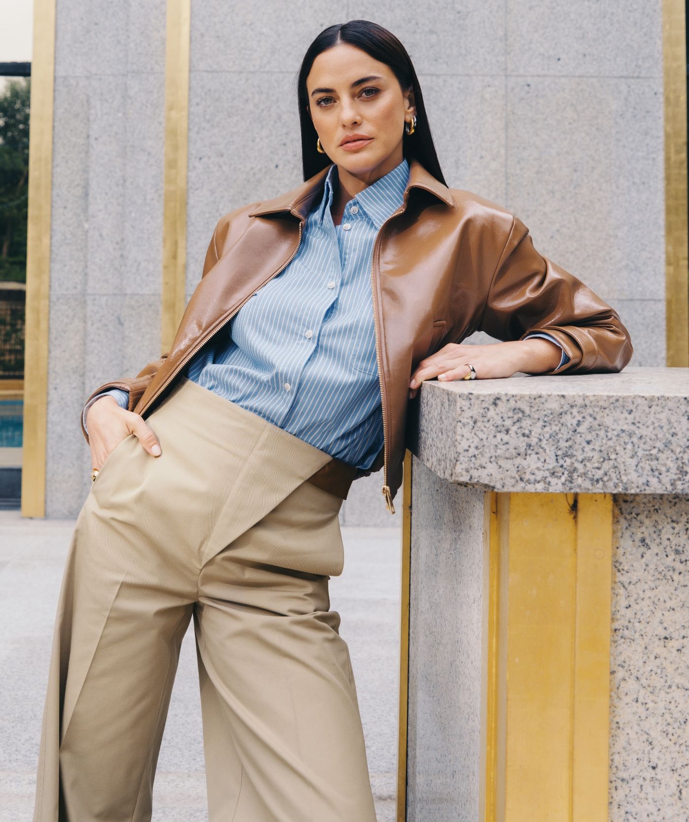 Model draagt de lichtblauw wit gestreepte blouse samen met een lak look jasje op een beige pantalon.  