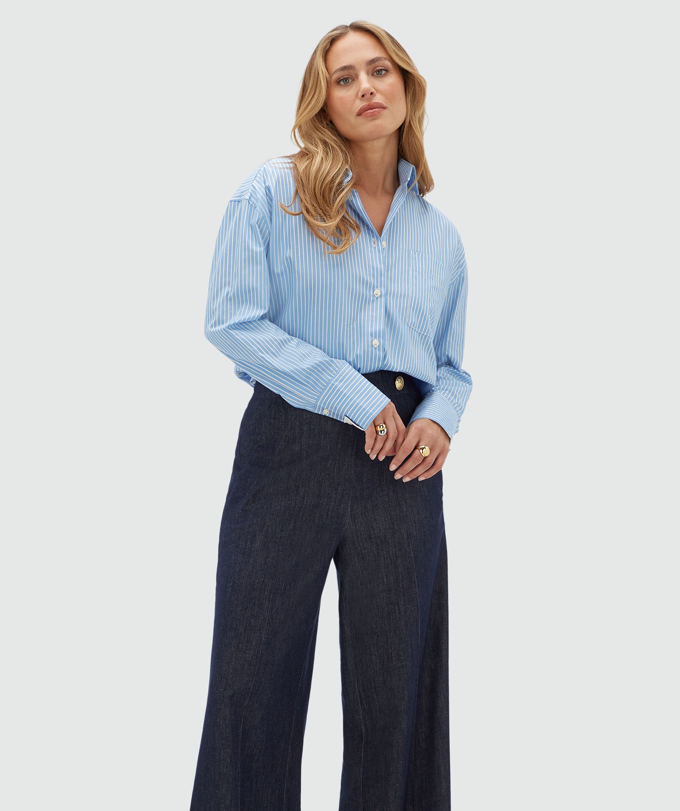 Model draagt een lichtblauw met wit gestreepte blouse gecombineerd met een donkerblauwe pantalon. 