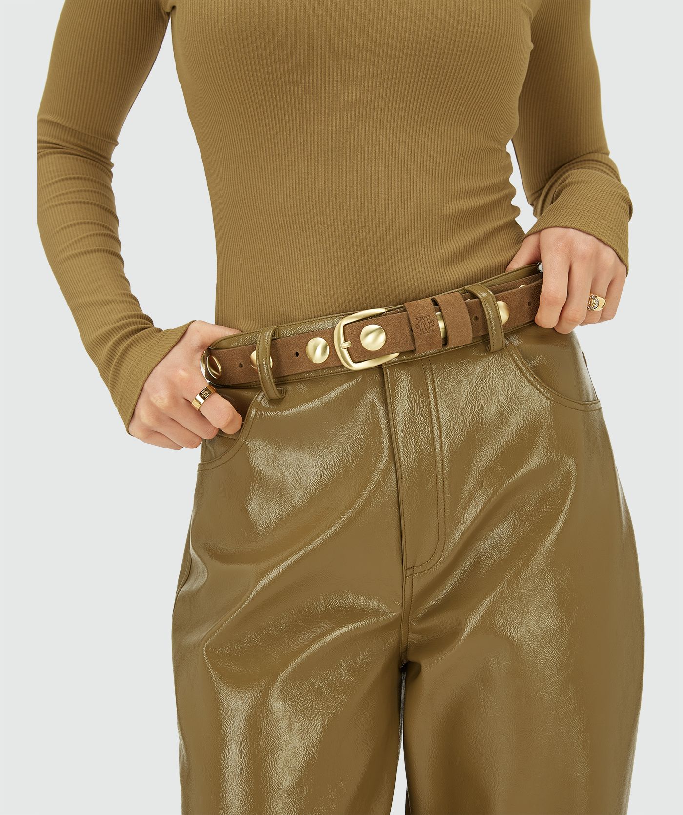 Model draagt een bruine suède riem met goudkleurige ronde studs en een goudkleurige gesp, afgewerkt met een subtiel JV-monogram logo bij de lus.