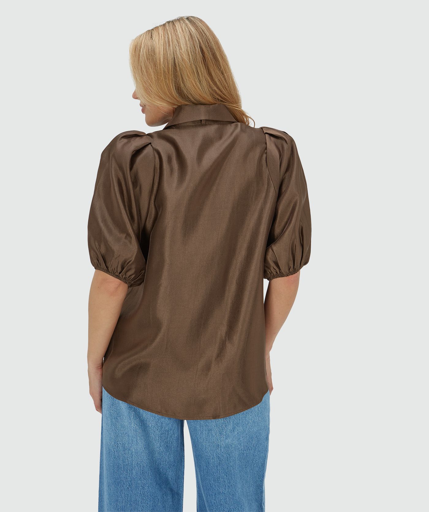 Achteraanzicht van het model in een regular fit blouse van een glanzende bruine stof, afgewerkt met een strikdetail bij de halslijn