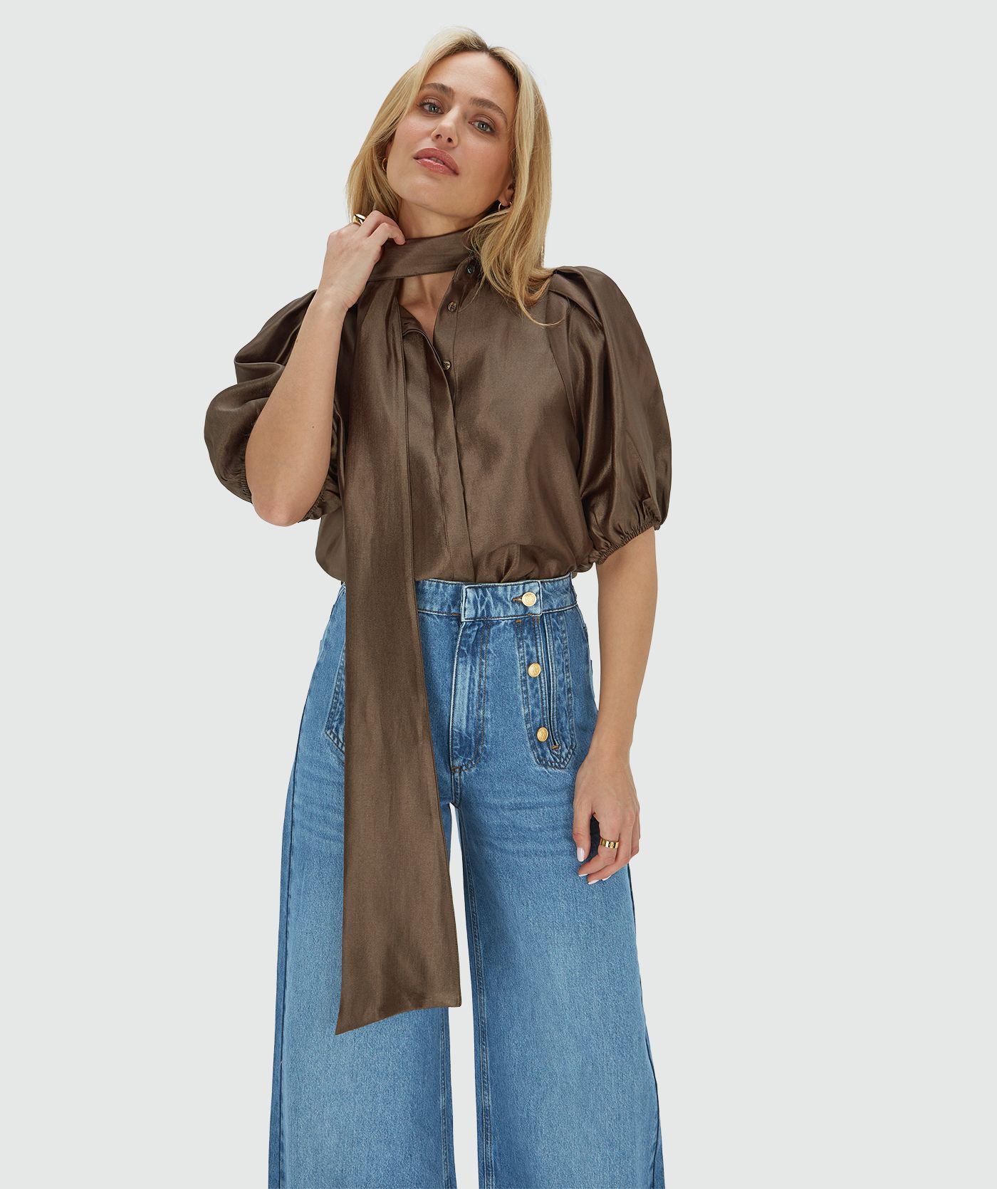 Het model draagt een regular fit blouse van een glanzende bruine stof, afgewerkt met een strikdetail bij de halslijn