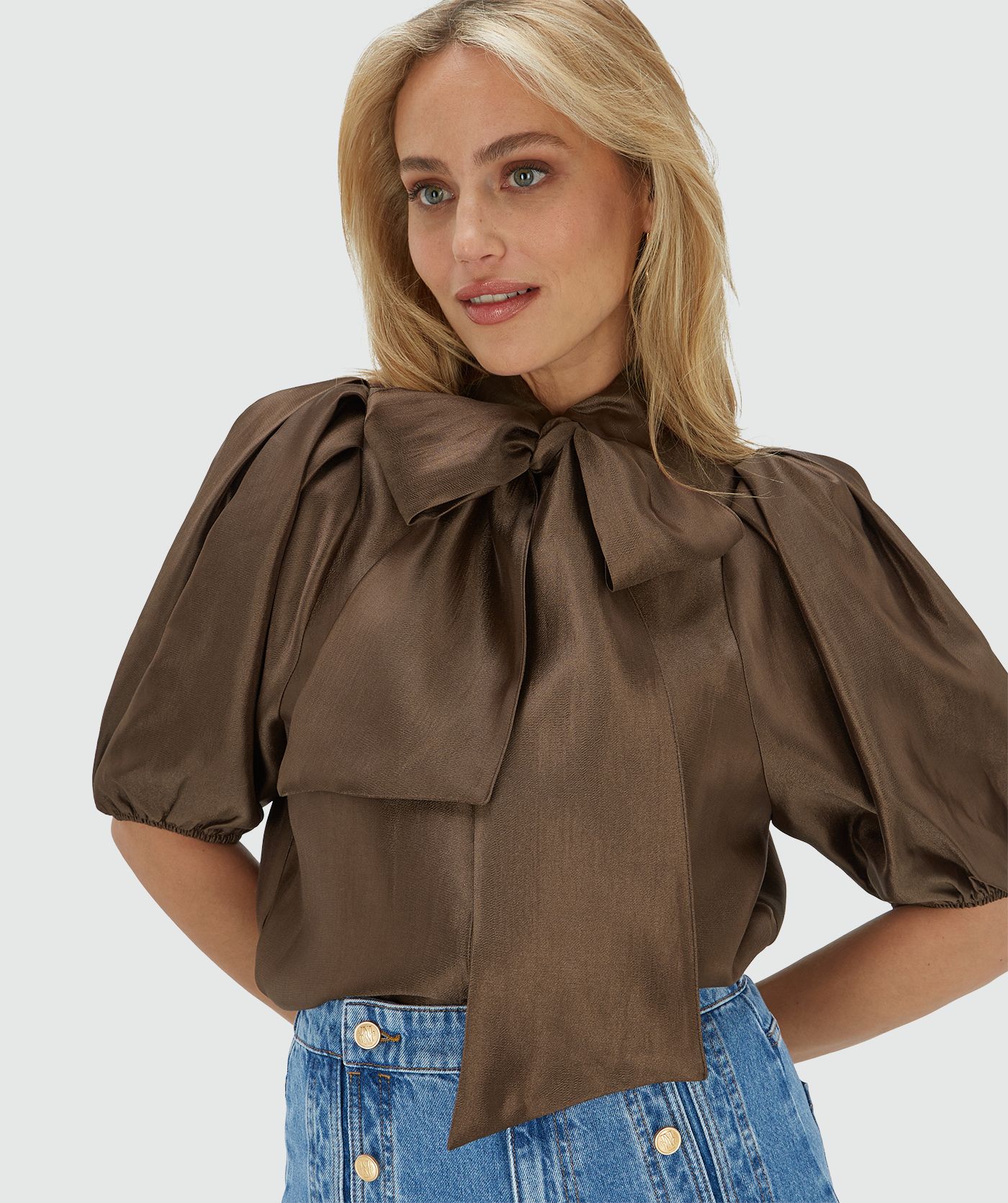 Het model draagt een regular fit blouse van een glanzende bruine stof, afgewerkt met een strikdetail bij de halslijn