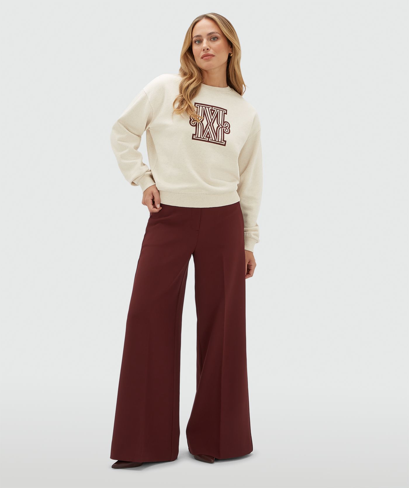 Model draagt een beige sweater met een donkerrood logo gecombineerd op een donkerrode pantalon.