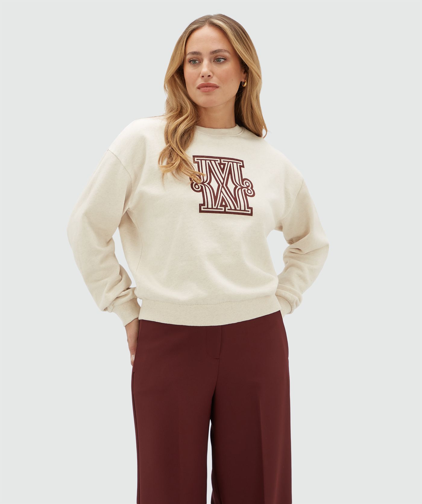 Model draagt een beige sweater met een donkerrood logo gecombineerd op een donkerrode pantalon.