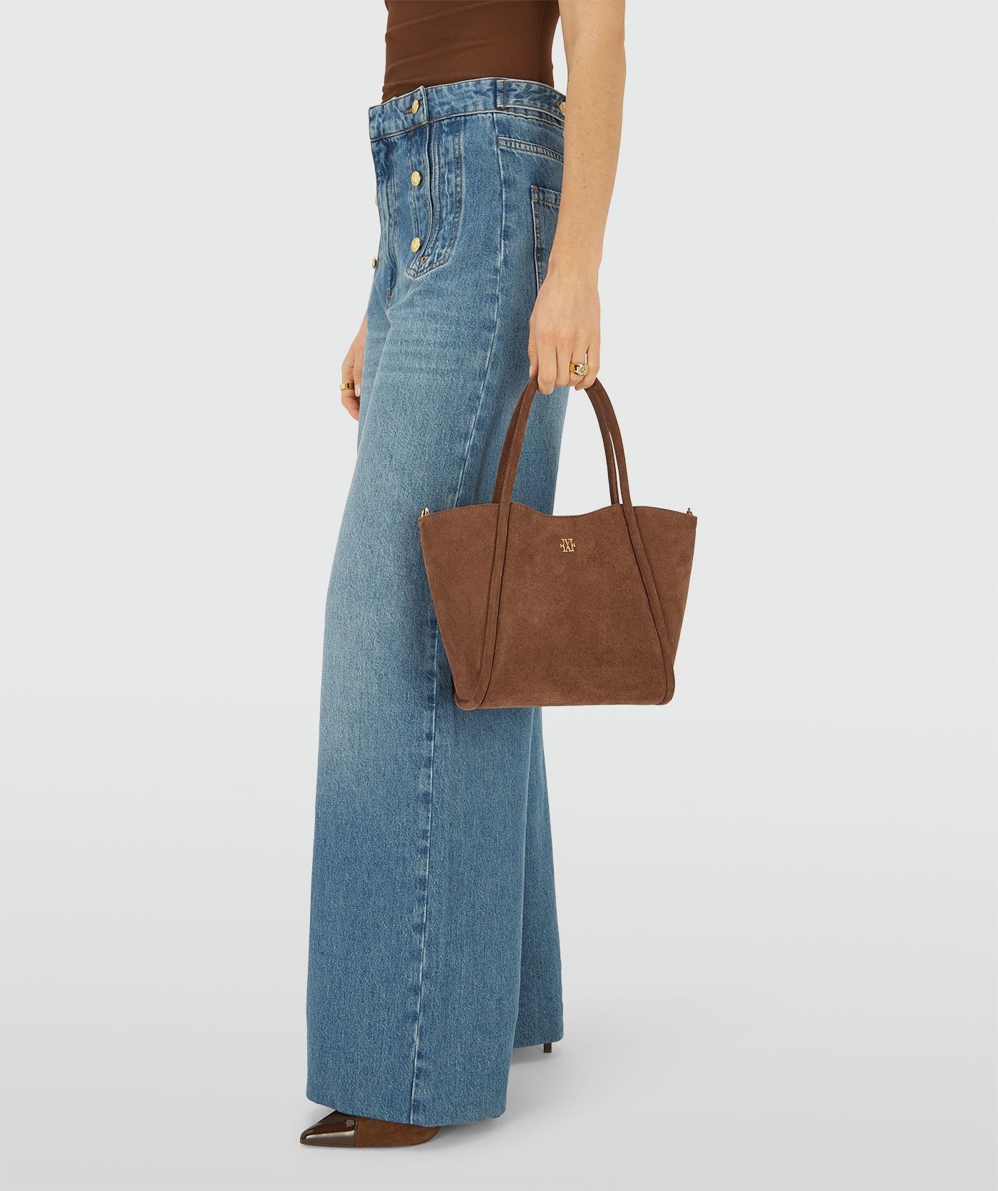 Model draagt een bruine suède handtas in trapezevorm met lange afneembare schouderhengsels afgewerkt met een goudkleurig JV-logo aan de voorzijde.