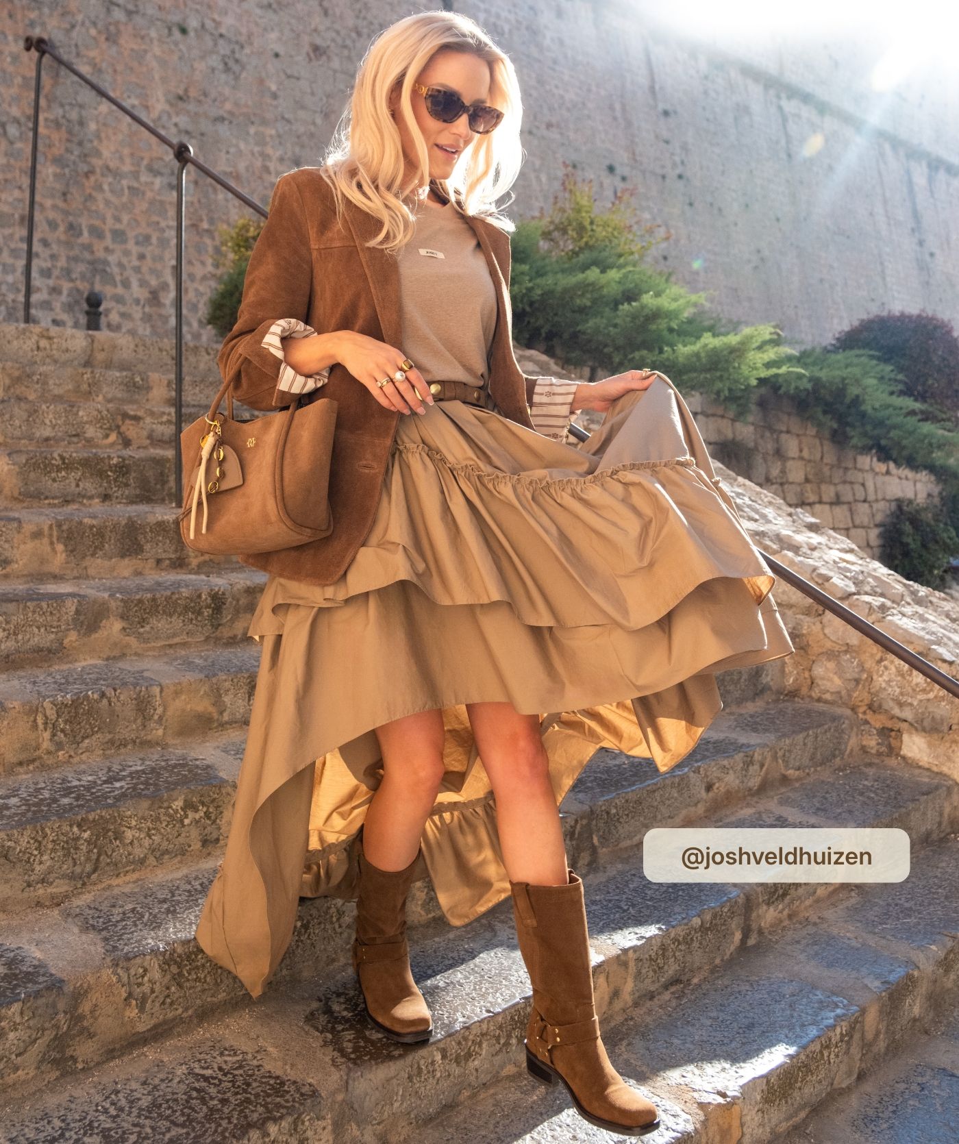 Josh Veldhuizen draagt een bruine suède handtas in trapezevorm met lange afneembare schouderhengsels afgewerkt met een goudkleurig JV-logo aan de voorzijde.