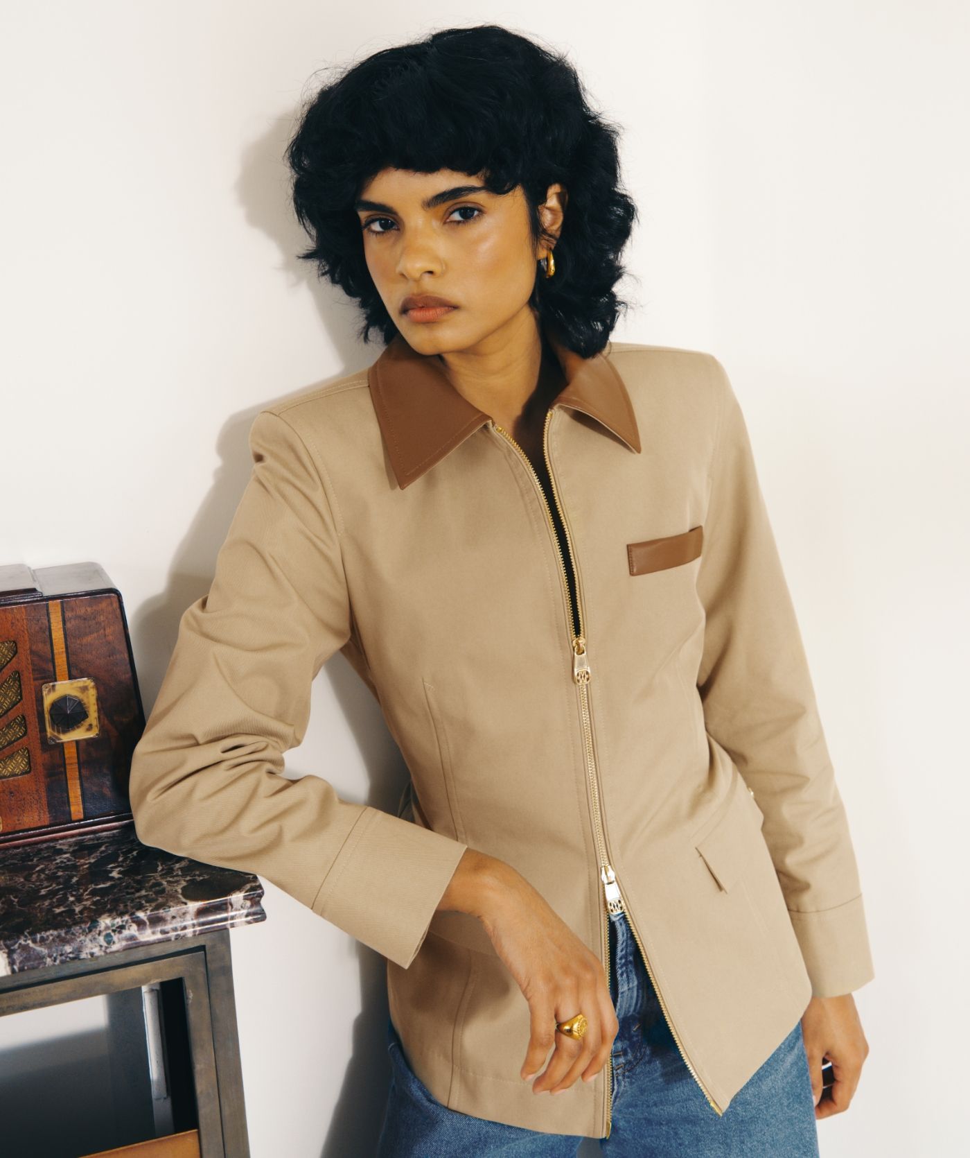 Model draagt het beige jasje met bruine leren details op een jeans en leunt tegen een kastje aan.