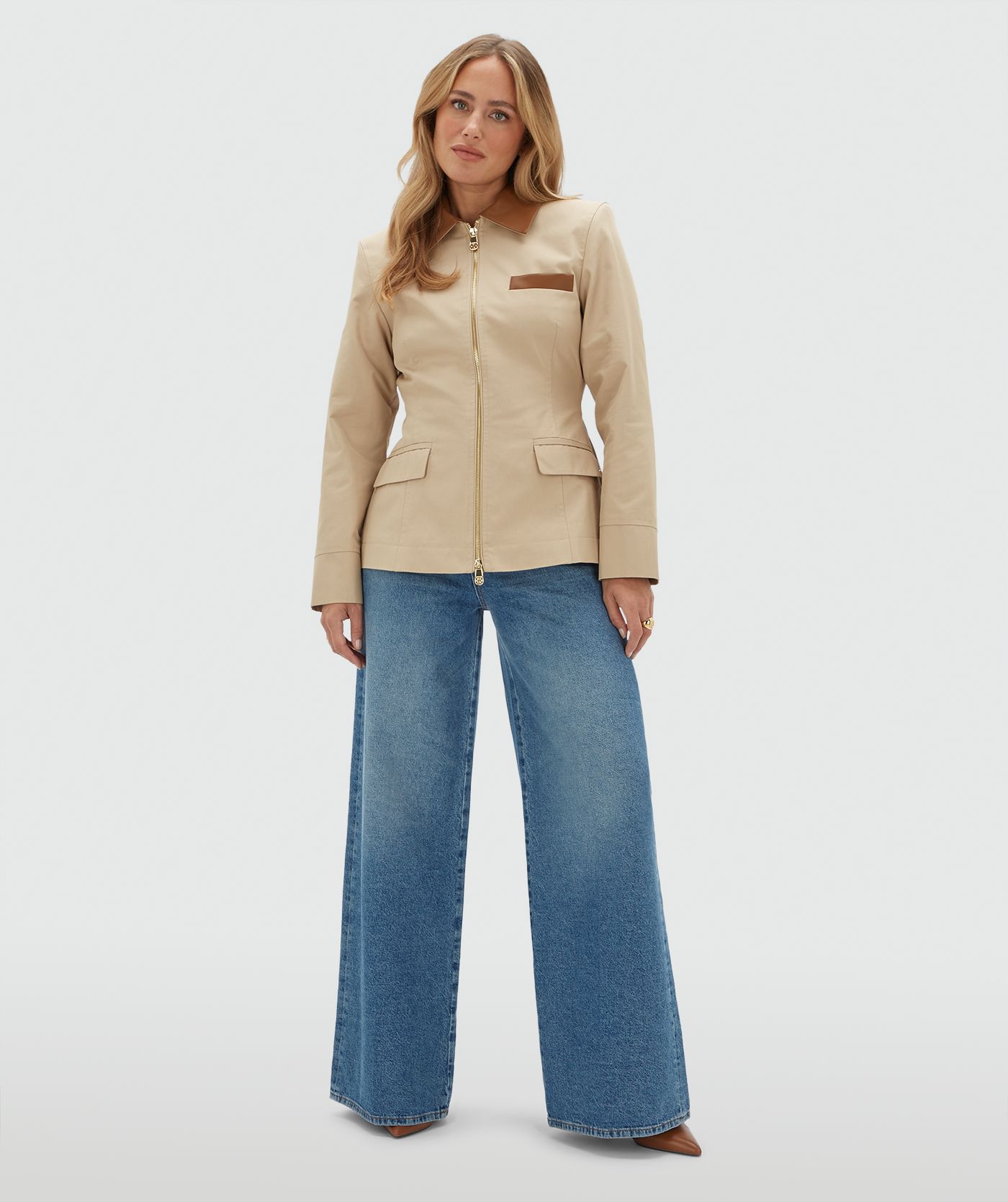 Model draagt een beige jasje met een rit en heeft een leren kraag en een leren detail bij het borstzakje aan de voorkant. Het jasje is gecombineerd met een bruine zonnebril en een jeans.