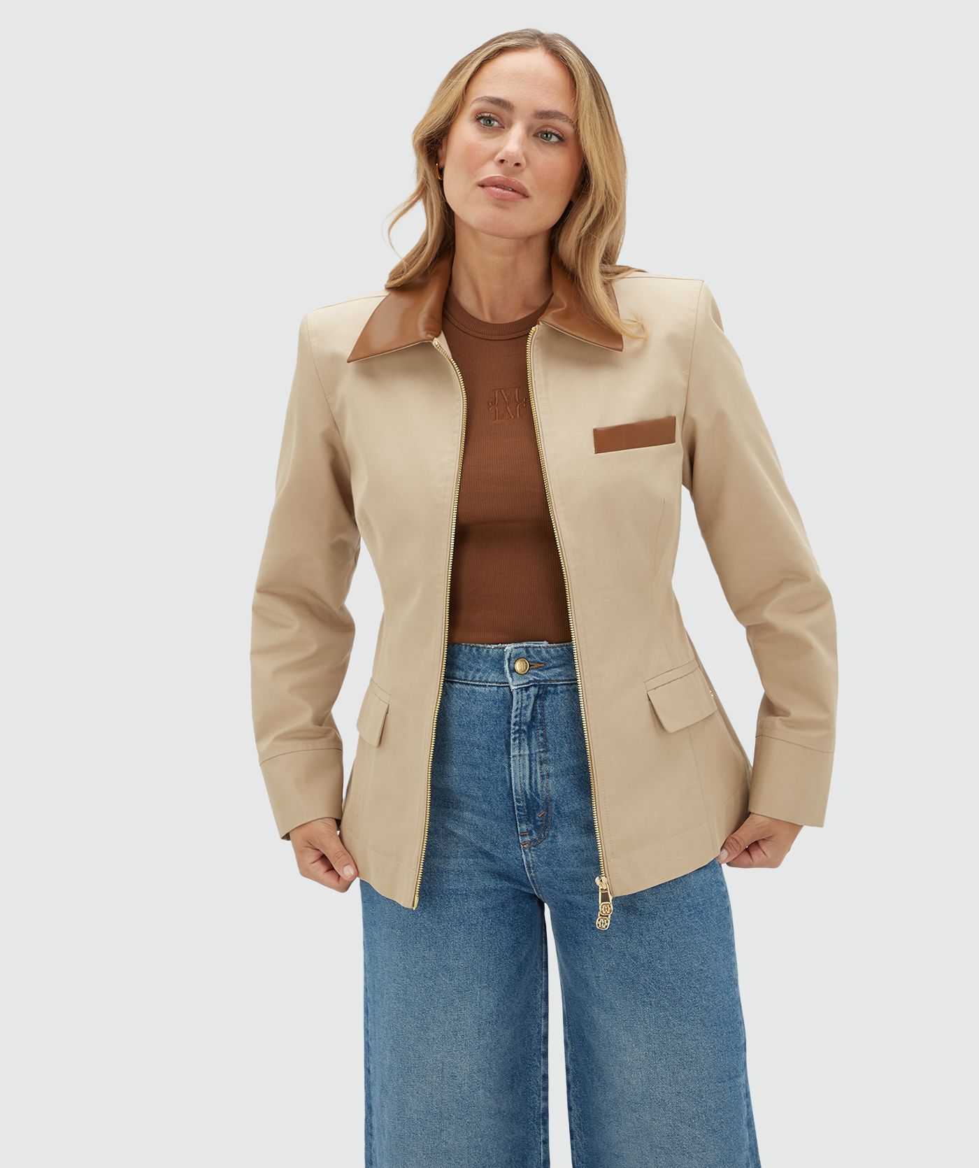 Model draagt een beige jasje met een rit en heeft een leren kraag en een leren detail bij het borstzakje aan de voorkant. Het jasje is gecombineerd met een bruine zonnebril en een jeans.