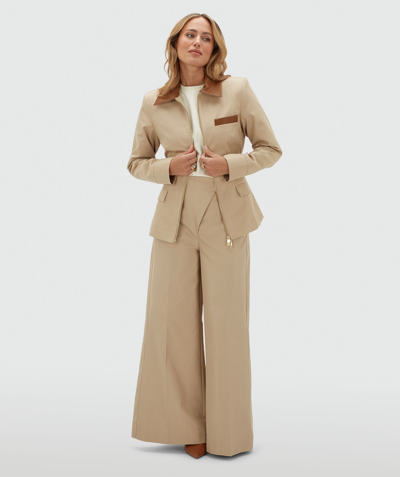 Model draagt een beige jasje met een rit en heeft een leren kraag en een leren detail bij het borstzakje aan de voorkant. Het jasje is gecombineerd met de bijpassende pantalon en draagt een wit t-shirt onder het jasje.