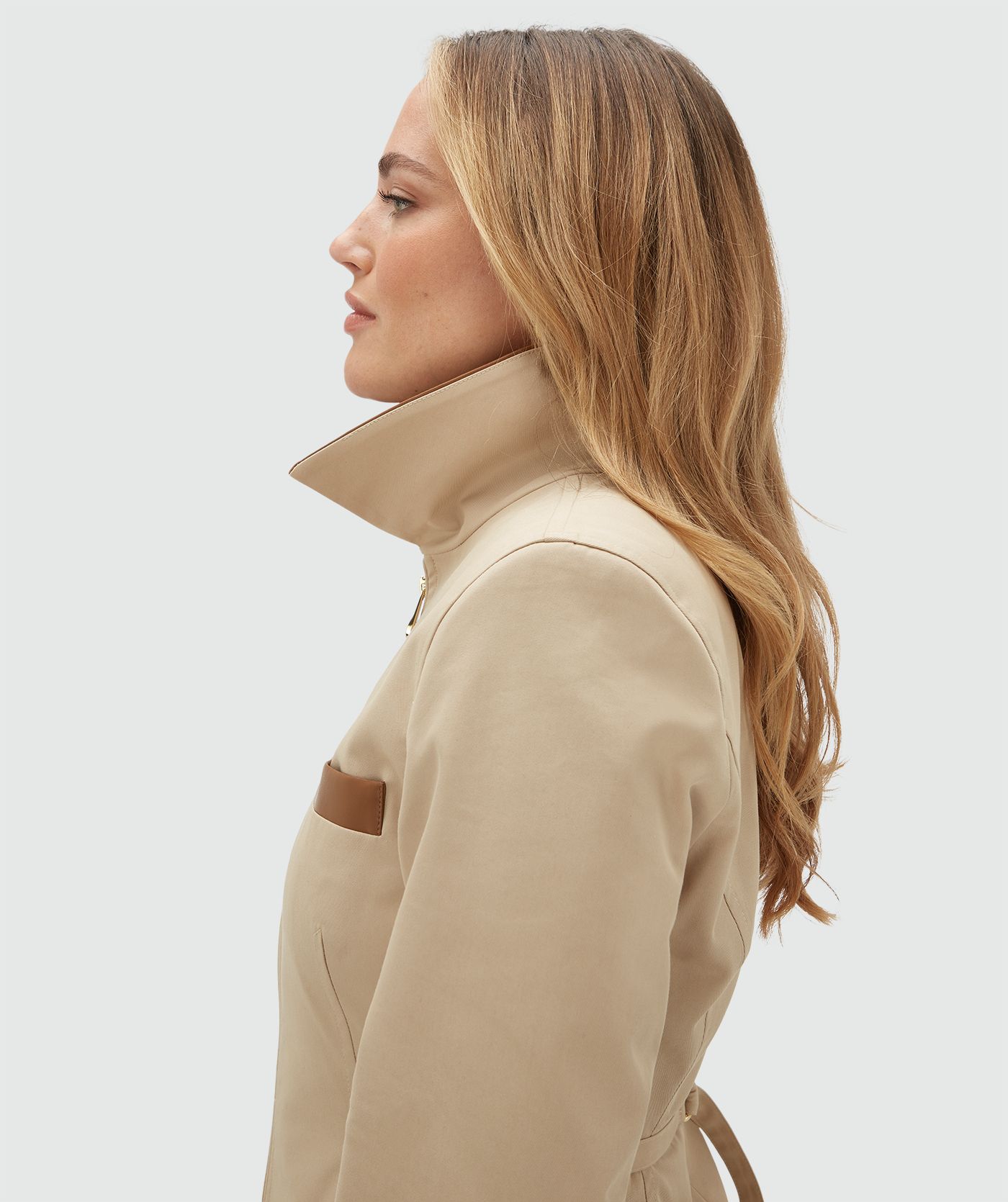 Model draagt een beige jasje met een rit en heeft een leren kraag en een leren detail bij het borstzakje aan de voorkant. Het jasje is gecombineerd met de bijpassende pantalon en draagt een wit t-shirt onder het jasje.