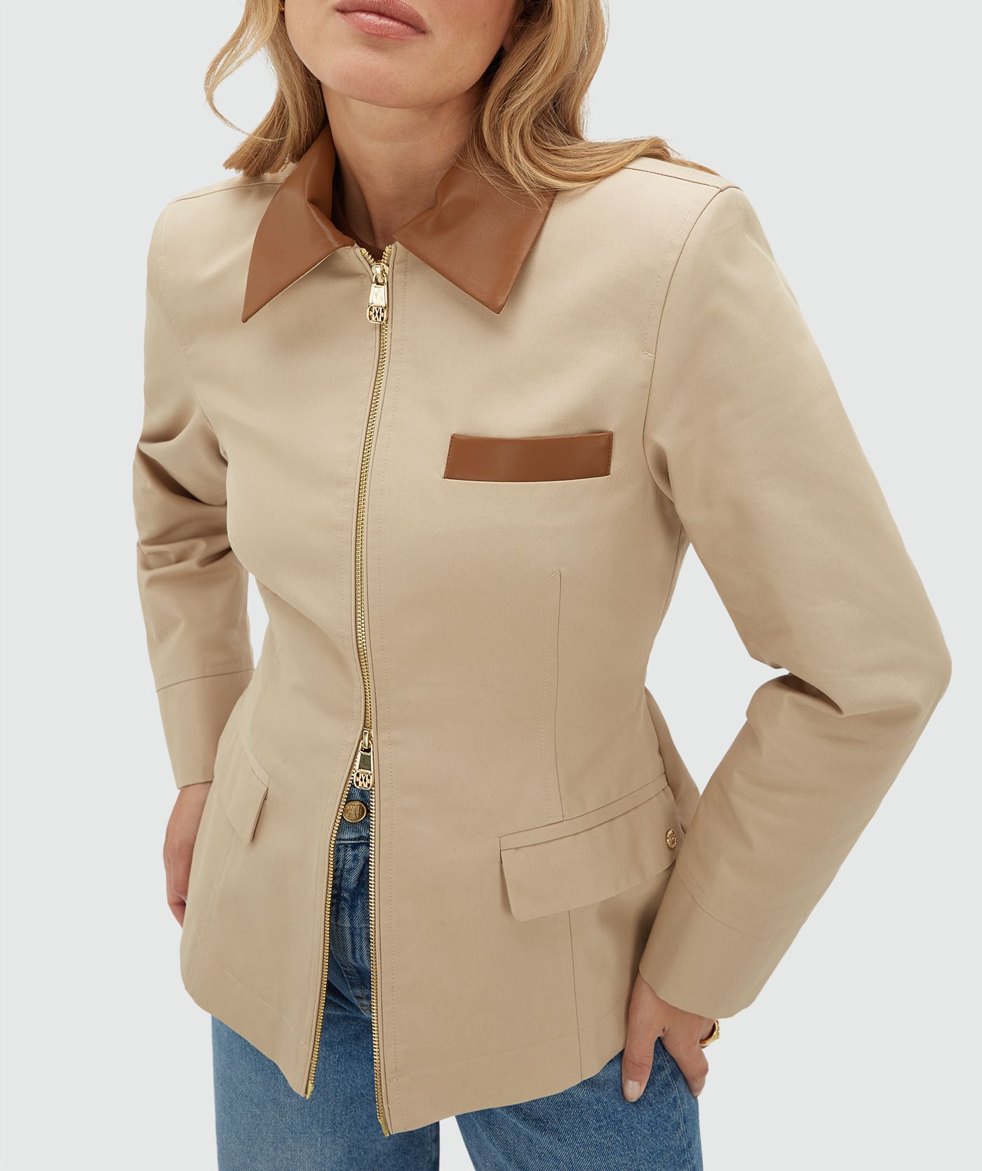 Model draagt een beige jasje met een rit en heeft een leren kraag en een leren detail bij het borstzakje aan de voorkant. Het jasje is gecombineerd met de bijpassende pantalon en draagt een wit t-shirt onder het jasje.