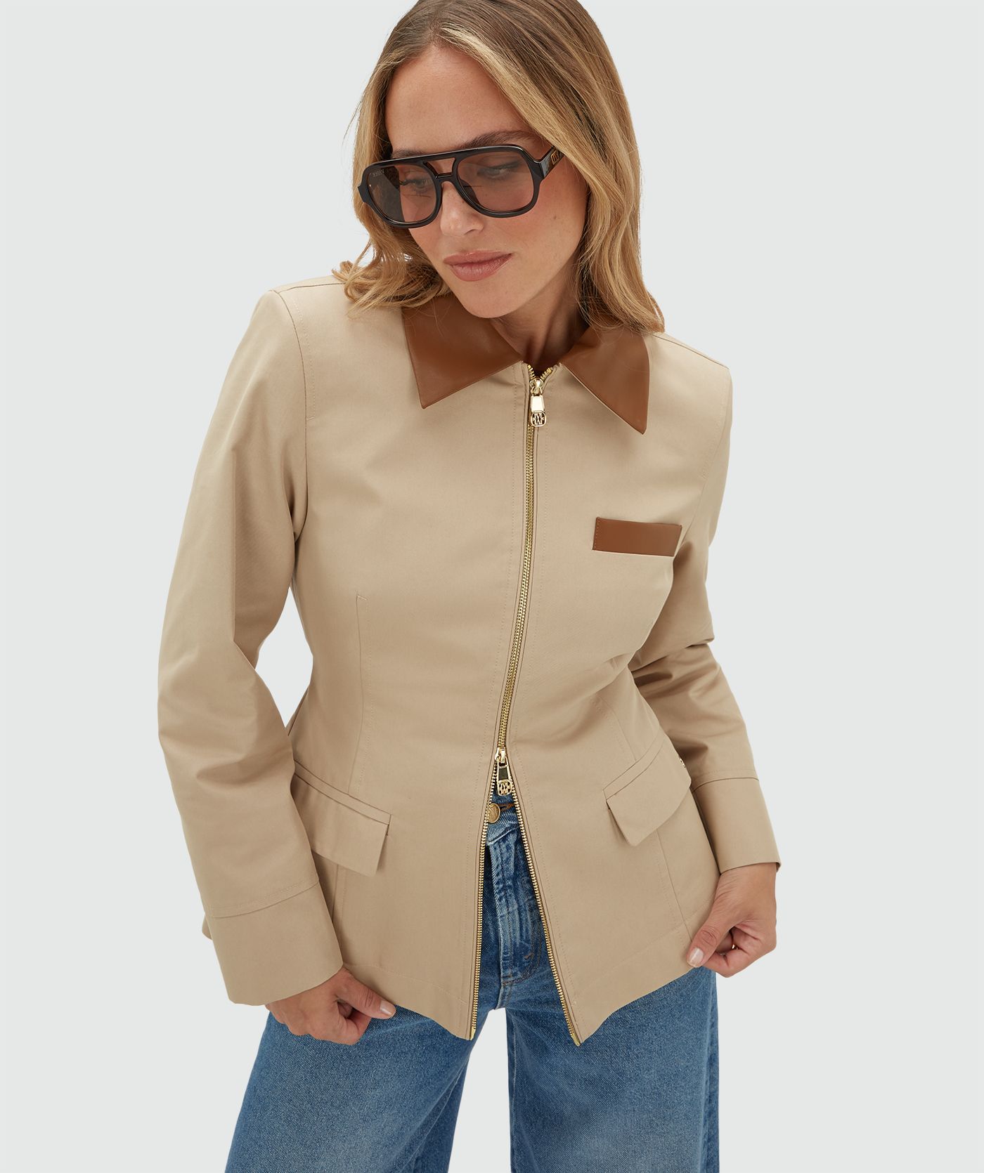 Model draagt een beige jasje met een rit en heeft een leren kraag en een leren detail bij het borstzakje aan de voorkant. Het jasje is gecombineerd met een bruine zonnebril en een jeans.