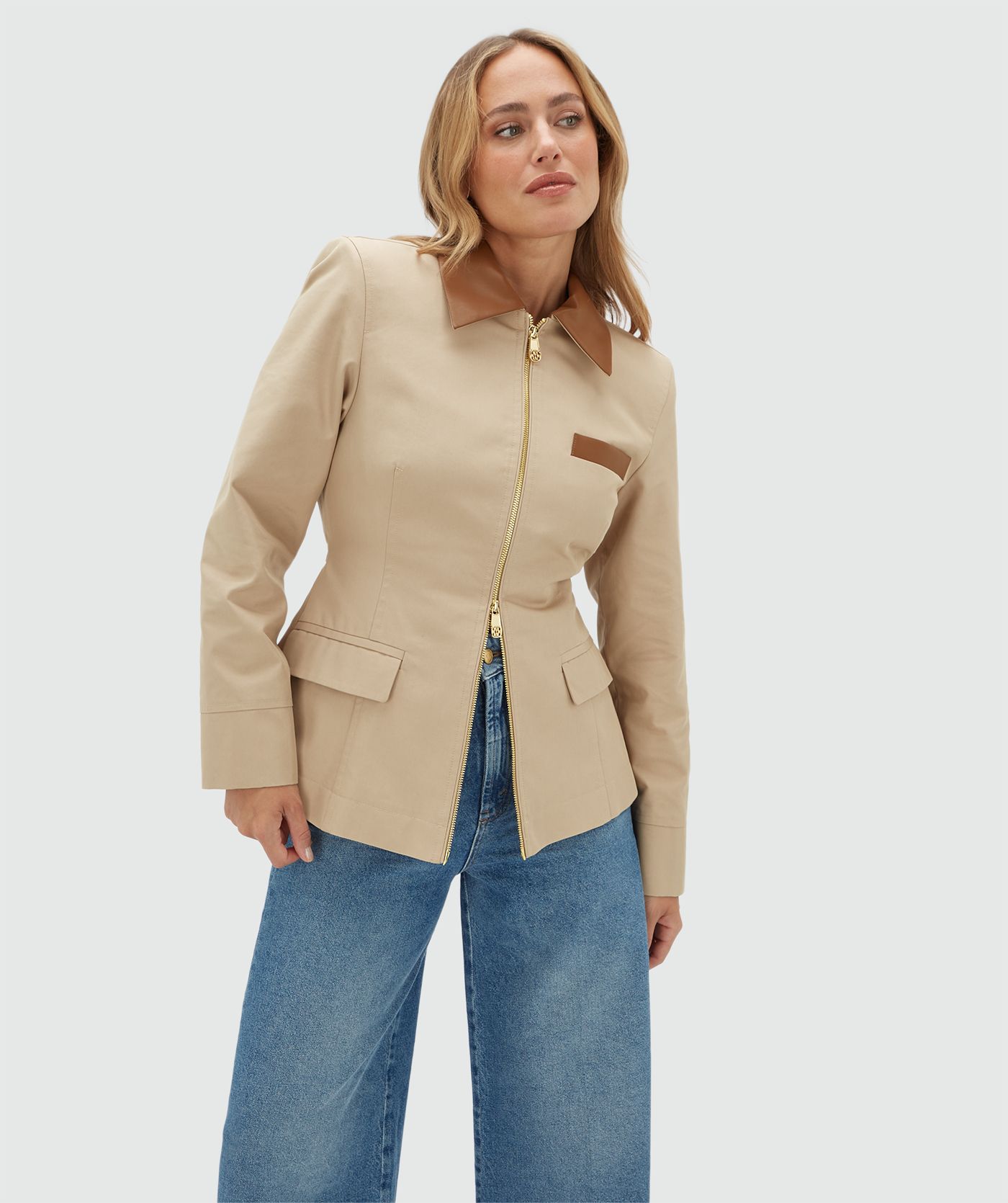 Model draagt een beige jasje met een rit en heeft een leren kraag en een leren detail bij het borstzakje aan de voorkant. Het jasje is gecombineerd met een bruine zonnebril en een jeans.