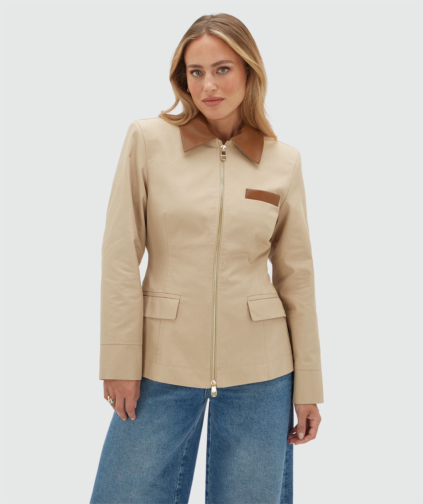 Model draagt een beige jasje met een rit en heeft een leren kraag en een leren detail bij het borstzakje aan de voorkant. Het jasje is gecombineerd met een bruine zonnebril en een jeans.