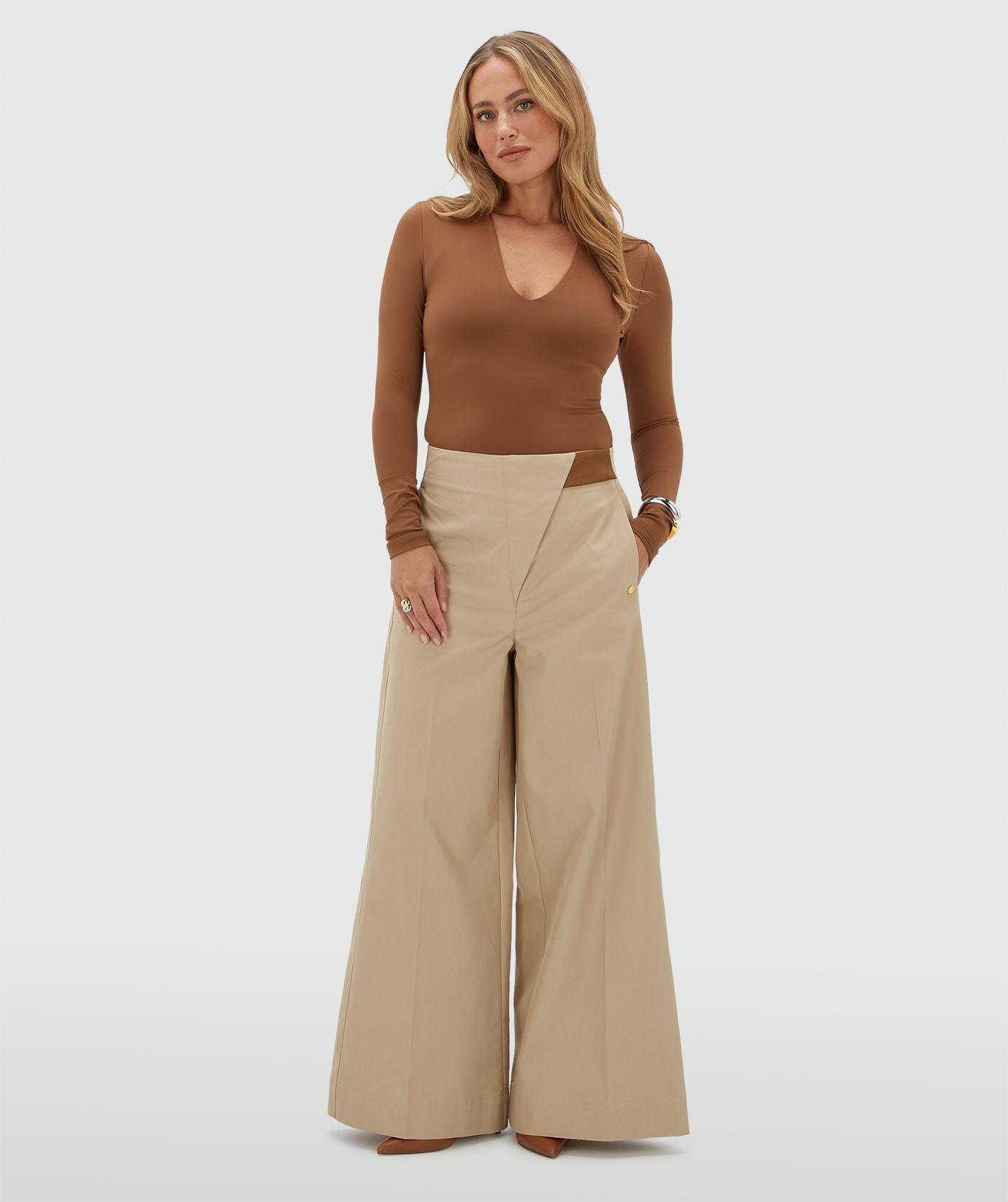 Model draagt een beige pantalon met aan de bovenkant een lichtbruin leren stukje waarmee je de broek sluit.