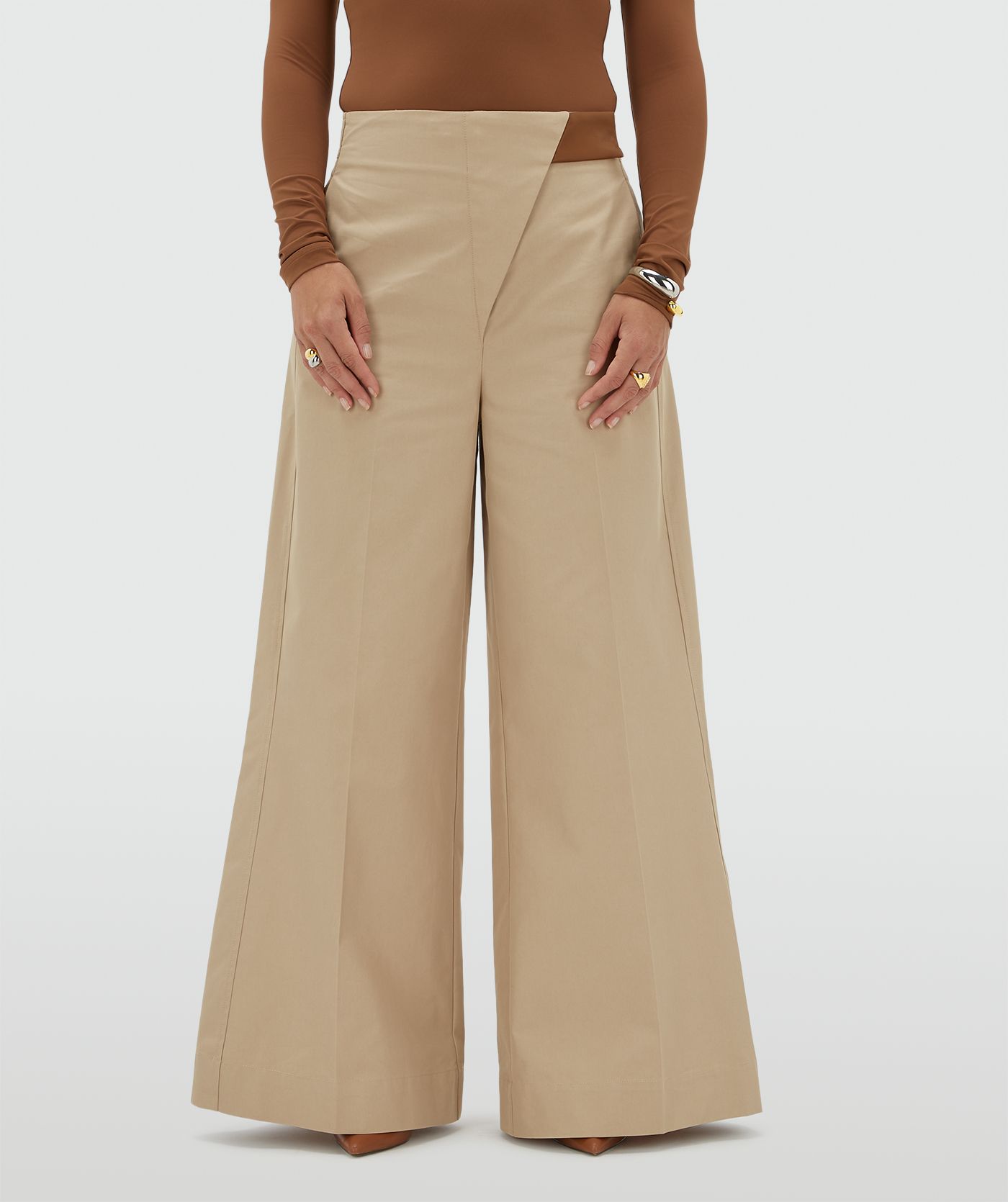 Model draagt een beige pantalon met aan de bovenkant een lichtbruin leren stukje waarmee je de broek sluit.