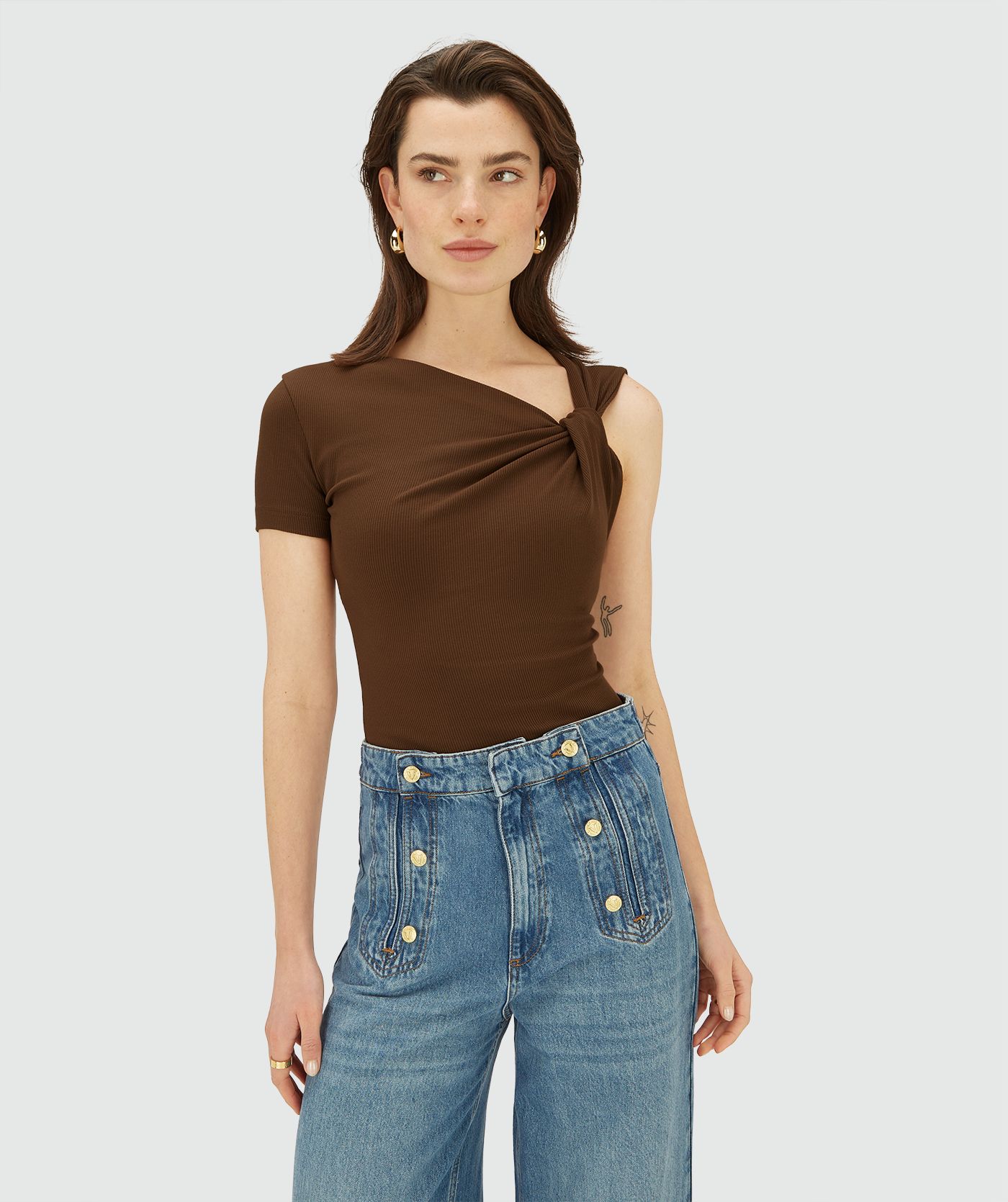 Model draagt een bruine, asymmetrische top met korte mouw, schuin halsdetail en knoopdetail op de schouder