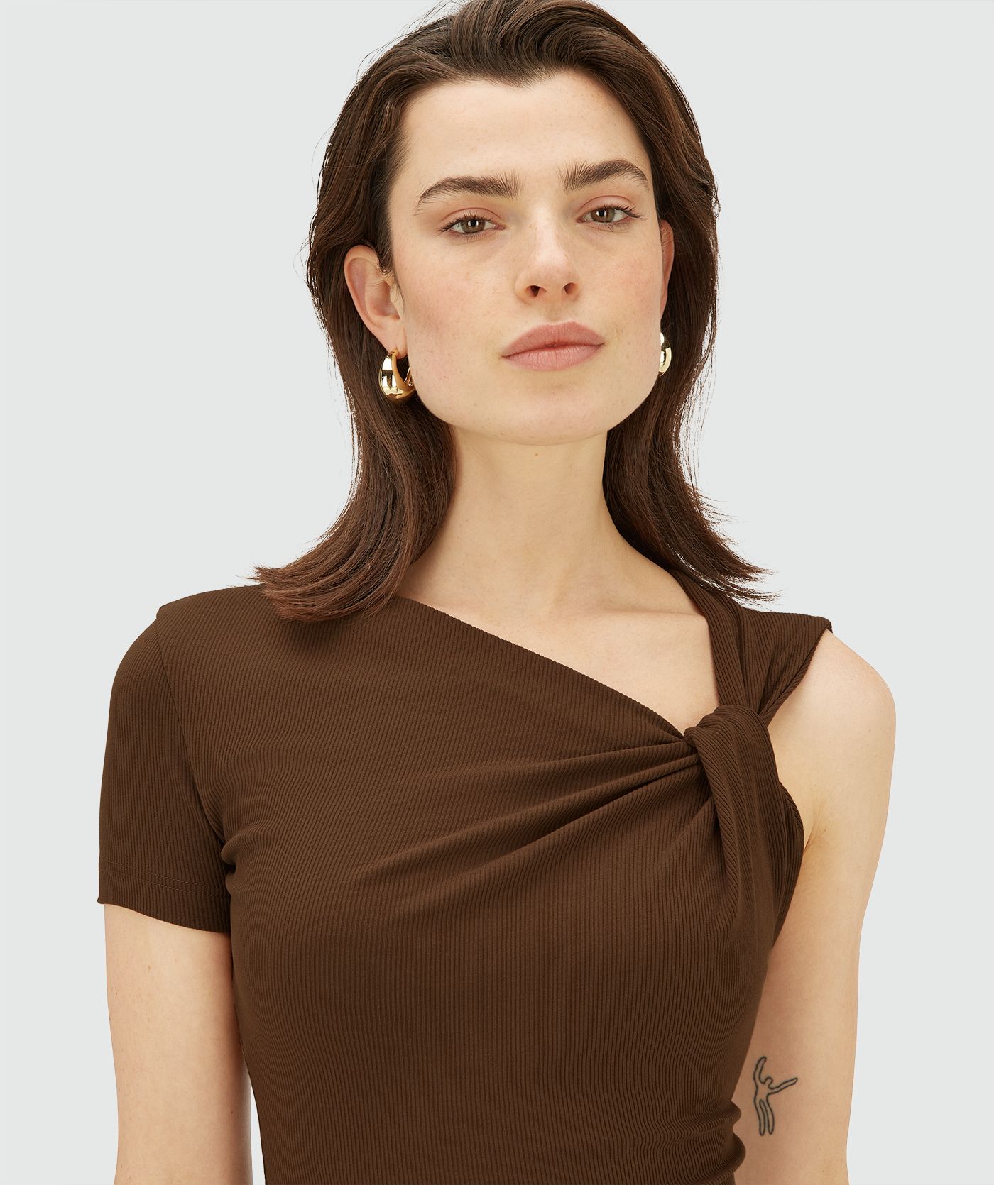 Model draagt een bruine, asymmetrische top met korte mouw, schuin halsdetail en knoopdetail op de schouder