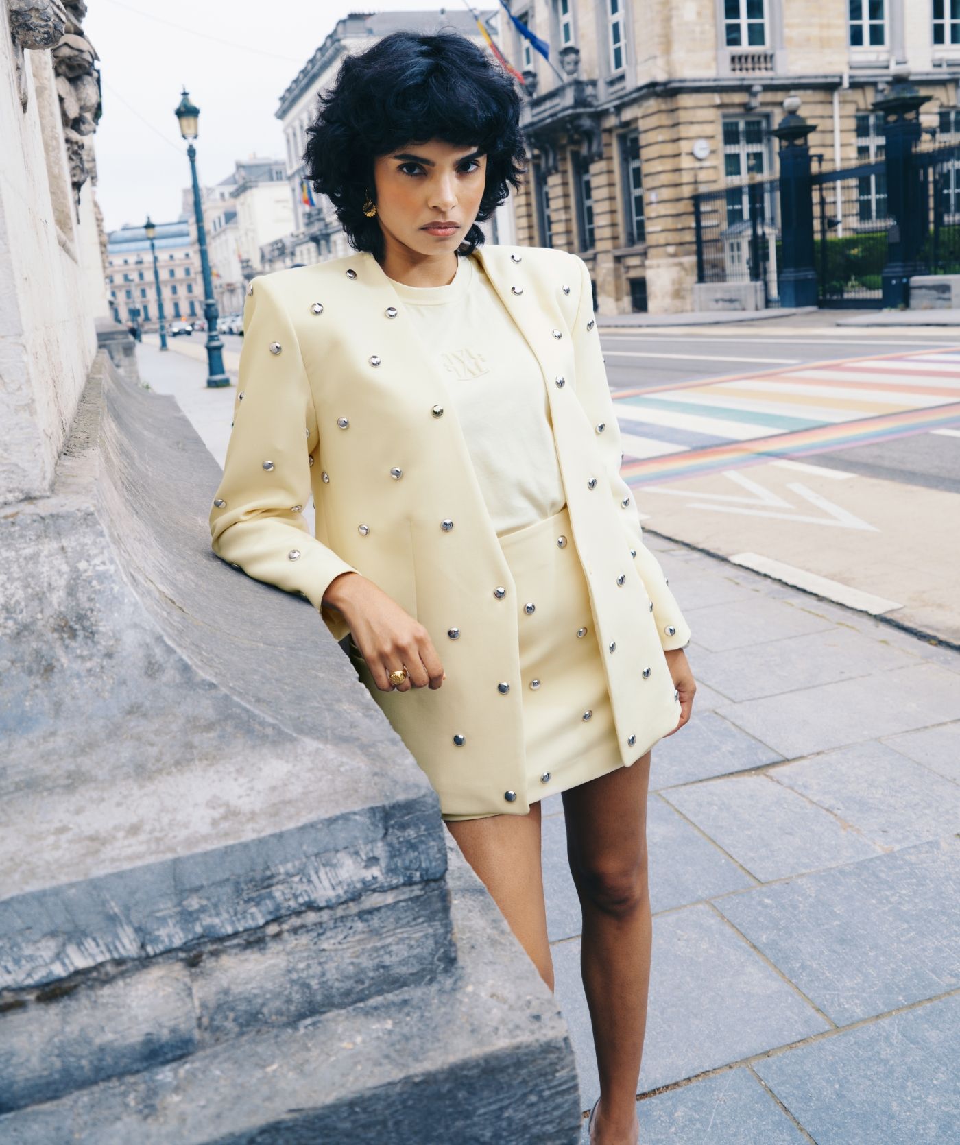 Model staat op straat en draagt lichtgele blazer met zilveren studs met het bijpassende rokje.